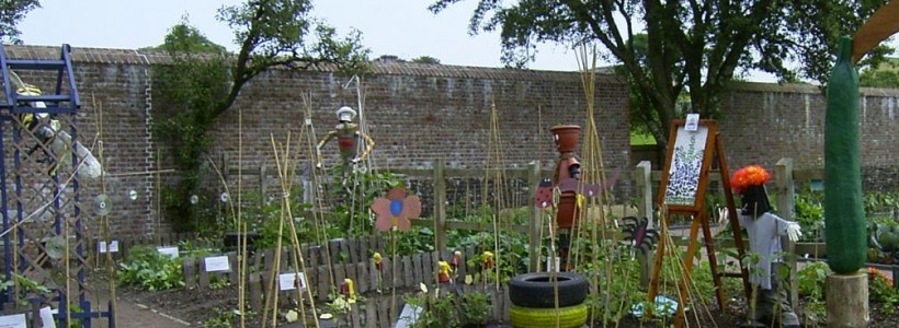A shot of a school garden with a path through the middle, things growing in tires, bean poles supporting plants and homemade scarecrows.